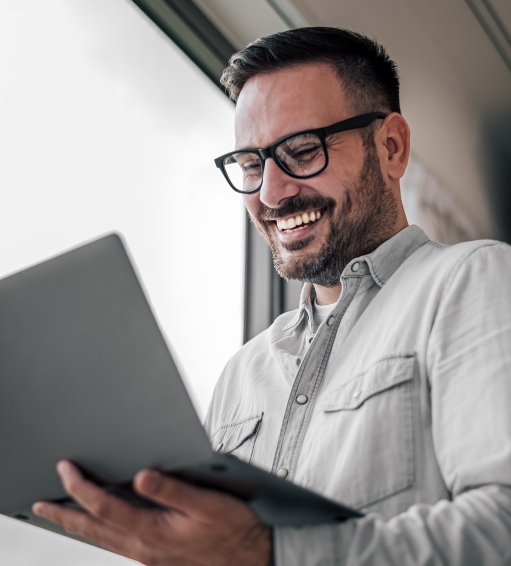 Smiling man wearing glasses looking at a laptop near a window.