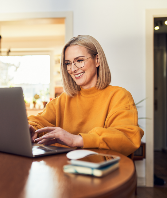 Smiling woman wearing glasses and a yellow sweater working on a laptop at a table.