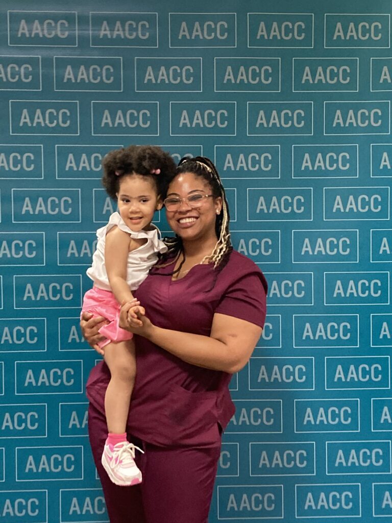 Woman smiling and holding a young girl in front of a blue AACC backdrop.