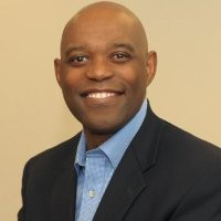 Person smiling in a dark blazer and blue shirt against a neutral background.