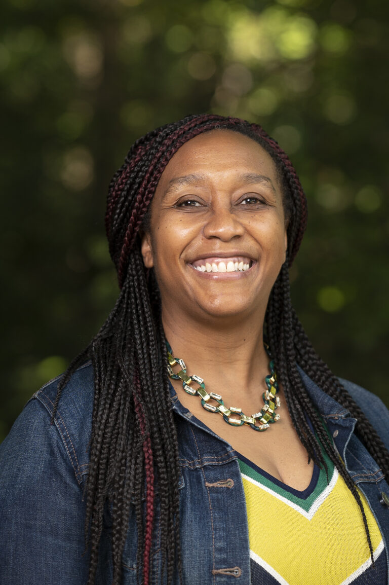 Person smiling outdoors wearing a denim jacket and colorful necklace, with trees softly blurred in the background.