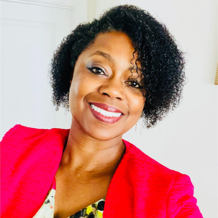 Person smiling in a red jacket and patterned top, standing indoors against a light background.