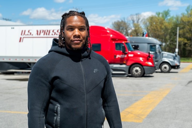 Person standing in a parking lot with red and gray semi-trucks in the background.