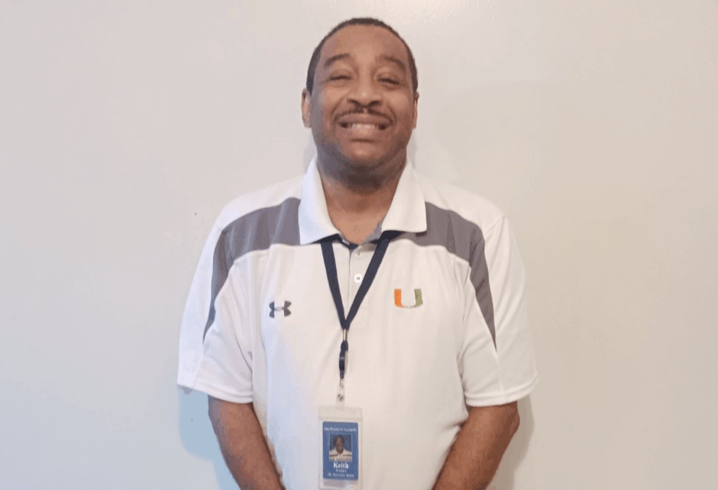 Man smiling while standing against a plain wall wearing a white polo shirt and an ID badge.